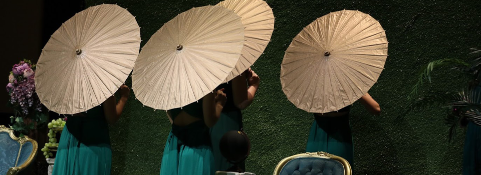 Singers stand in green dresses facing away from the audience with gold parasols open behind them.