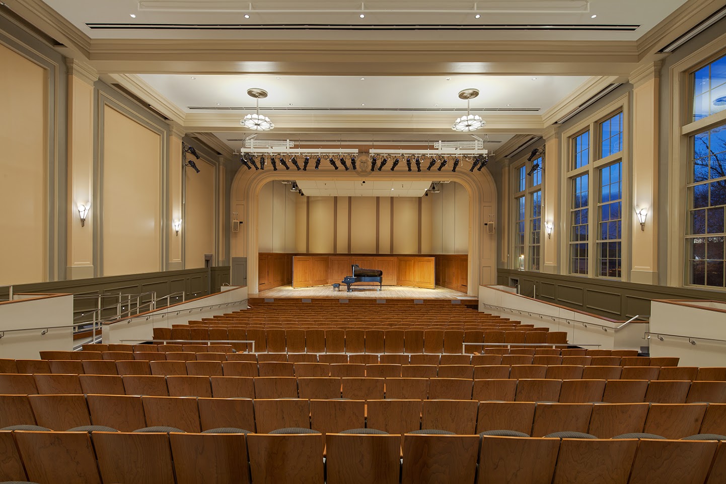 Moeser Auditorium, as seen from the back of the seats. The auditorium is empty, the lights are bright with a lone grand piano on the stage and the deep blue sky of twilight visible through the windows to the right.