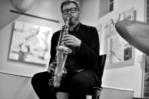 Brad Linde plays his saxophone sitting on a stool in an art gallery.