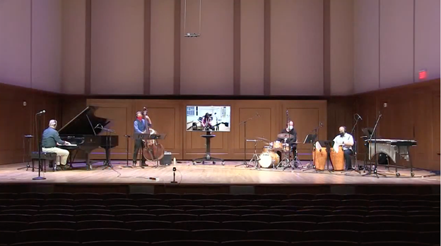 Jazz faculty perform on stage together, masked and socially distanced with saxophonist Barber playing along on a video screen from Hill Hall 103.