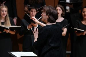 Susan Klebanow seen from over her left shoulder, conducting the Chamber Singers, in concert and smiling.