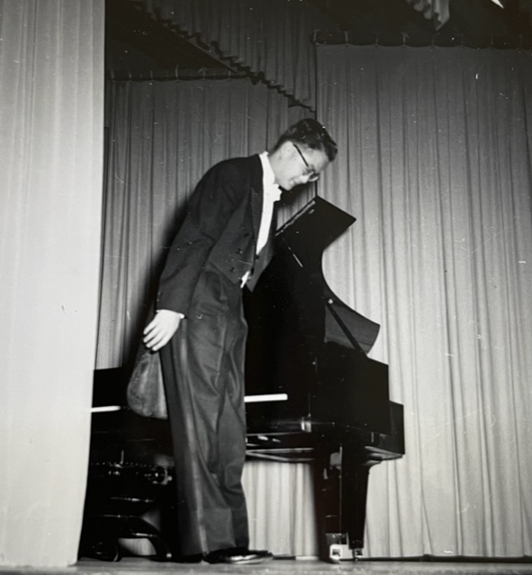 Francis Whang, in a tuxedo, bows to an audiences while next to a grand piano on a stage.