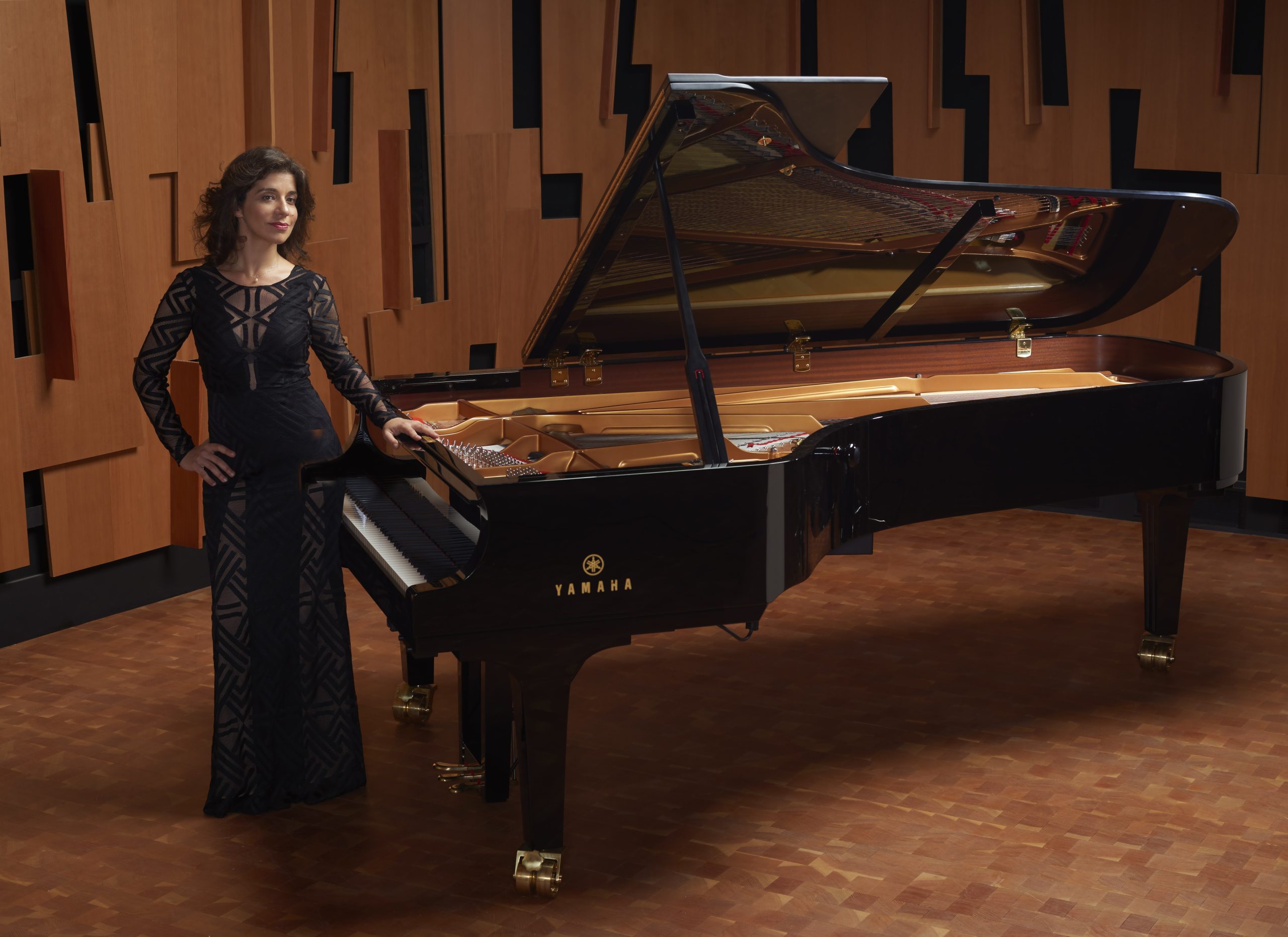 Inna Faliks stands next to a grand piano with her left hand resting on the instrument just above the keys. She wears a full-length, form-fitting, black gown.