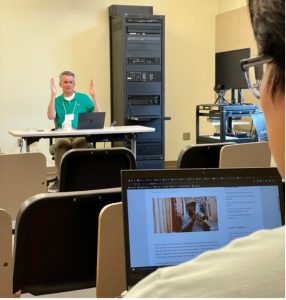 Andy Bechtel sits at the front of the class room, both hands raised to each side of this face. He is seen from the perspective of looking over a student's shoulder, their open laptop visible with a jazz article pulled up on their screen.