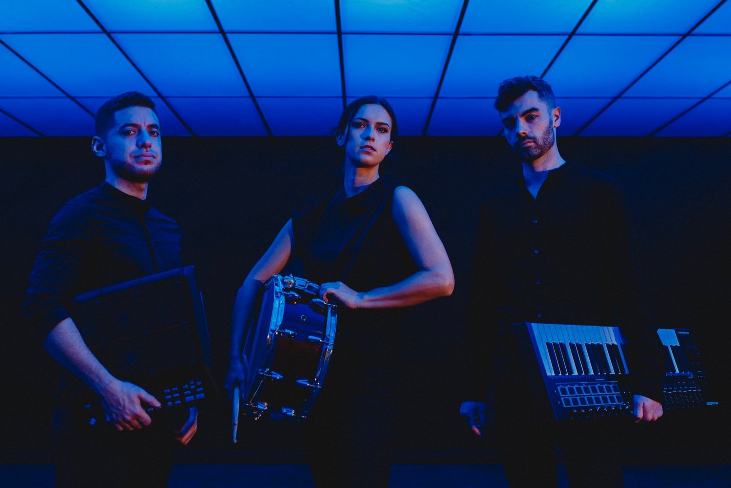 Pathos Trio stands dimly lit by blue lights holding their instruments.