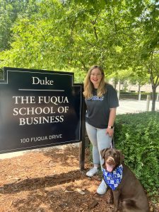 Courtney Hedgecock stands with her dog by the sign for the Duke Fuqua School of Business.